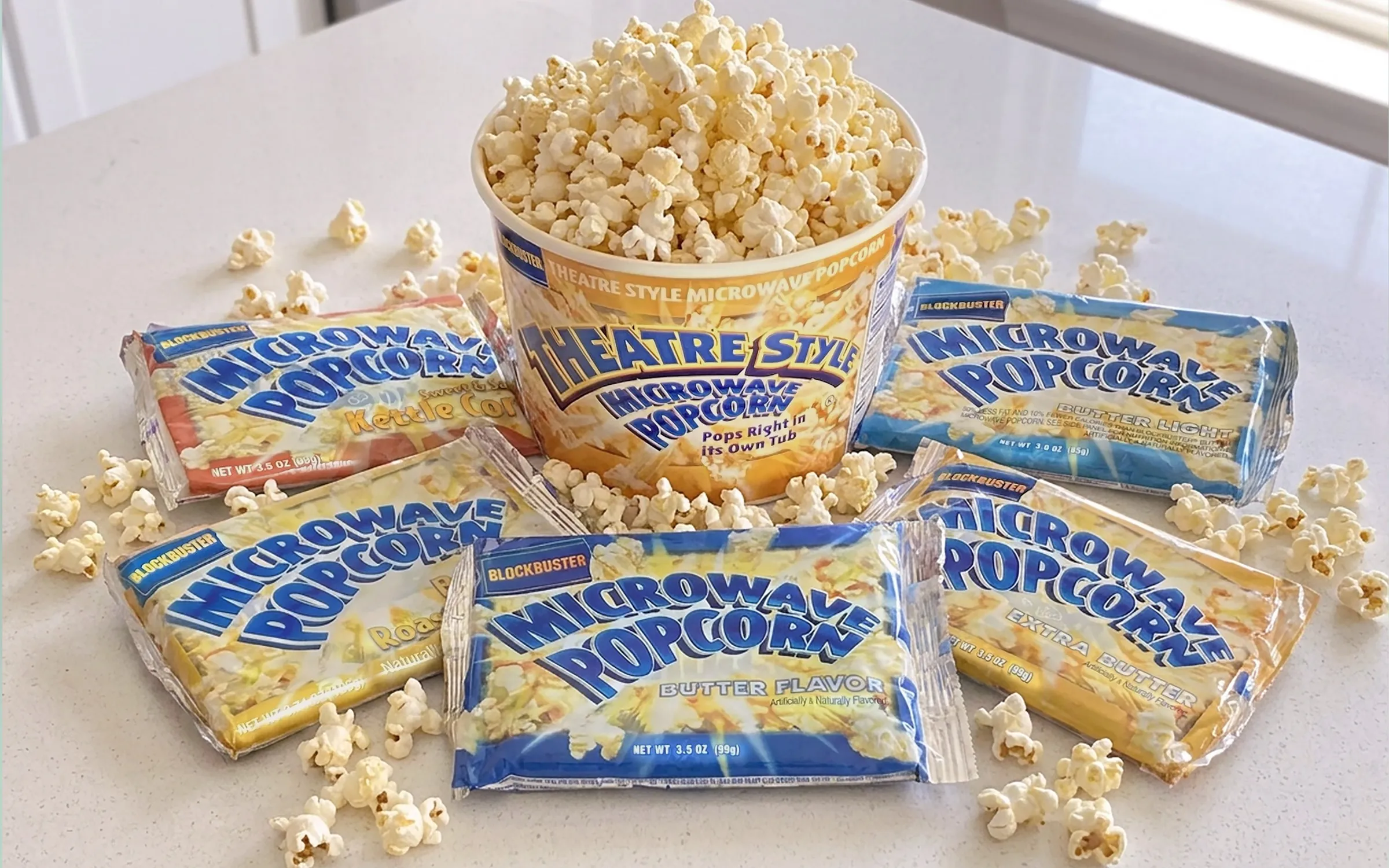 The Blockbuster microwave popcorn line laid out on a counter: a Theatre Style tub filled with popcorn, ringed by Kettle Corn, Butter Light, Butter Flavor, Extra Butter, and Natural packets.