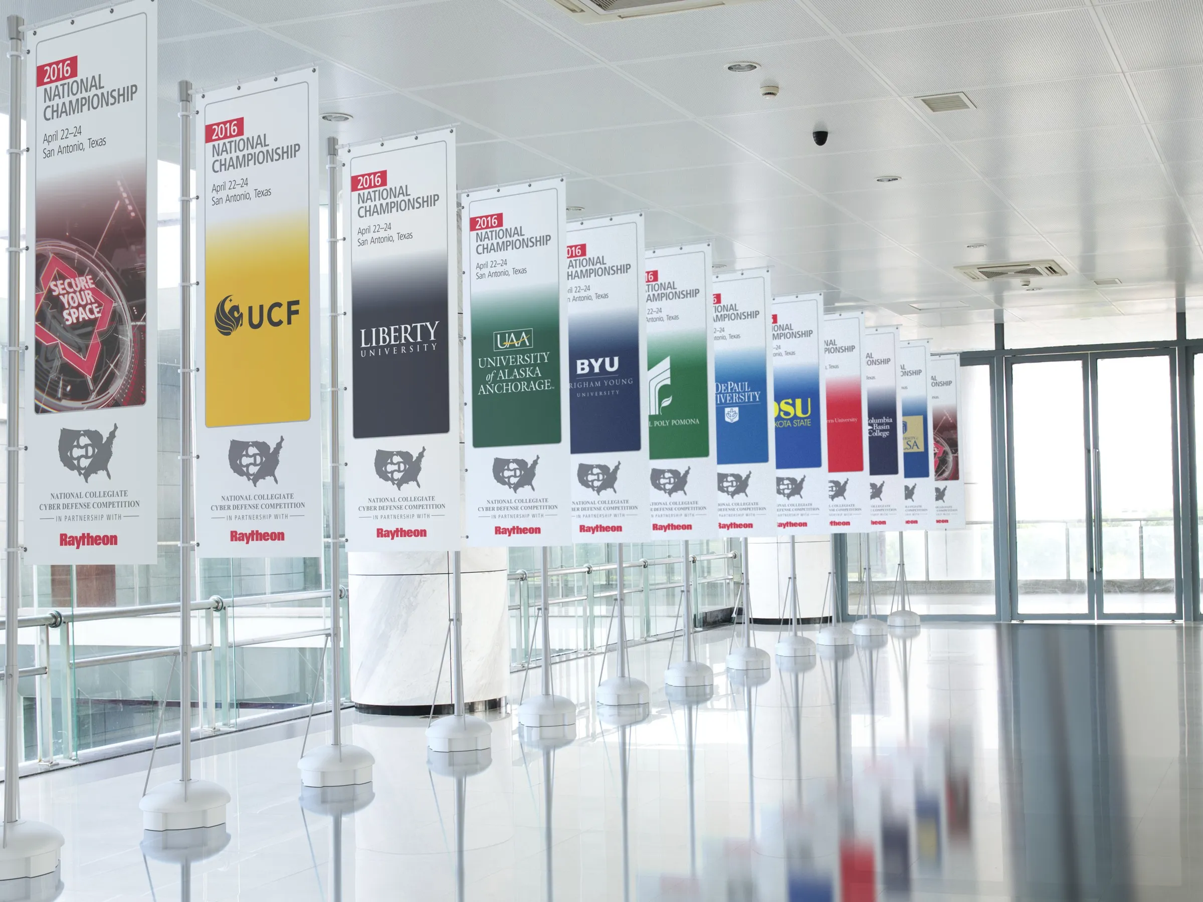 A long row of competition banners in a glass concourse, the first carrying the Secure Your Space mark and the rest the crests of competing universities.